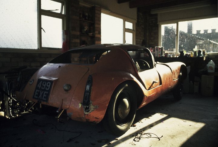 Another mystery car - Page 11 - Classic Cars and Yesterday's Heroes - PistonHeads - The image depicts a vintage, orange-colored vehicle with visible rust and damage. It has an open top and a classic design, reminiscent of older sports or racing cars. The car appears to be in a garage or indoor storage area, with natural light illuminating the interior. There are various items scattered throughout the space, suggesting a workspace or hobby area for vehicle restoration or repair.