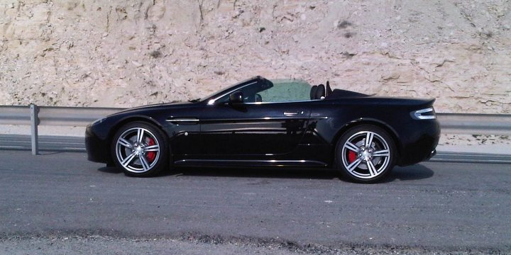 Hello from Cyprus! - Page 1 - Aston Martin - PistonHeads - The image portrays a black convertible car parked on the side of a road with a stone wall in the background. The vehicle is partially visible, allowing us to see the front part of the car, including the rims of the wheels and the driver's side door. The car's top is down, revealing a sleek design. The scene suggests it might be a clear day given the visibility of the surroundings and the car's shiny exterior.