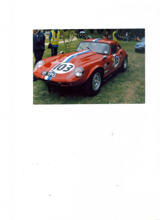 Pistonheads Palace Great Crystal Battles - The image features a striking red race car parked on grass with a greenish-brown undertone. The car is adorned with the number "103" in a prominent white color, suggesting a racing context. There are two people partially visible in the background, possibly spectators or team members associated with the car. The presence of another car, at a greater distance, subtly indicates that this could be a racing event or a trackday. The overall setting seems to be an outdoor event where motorsports are likely the main focus.