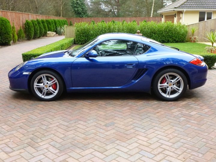 show us your toy - Page 107 - Porsche General - PistonHeads - The image features a sporty blue Porsche Cayman S parked on a brick driveway. The car is facing towards the right side of the photo, and it appears to be well-maintained with a shiny exterior. The driveway is bordered by a neatly trimmed hedge, and there are other structures and plants visible in the background, suggesting a residential area. The sky is overcast, casting a soft light on the scene. There's no visible text or distinctive branding in the image.