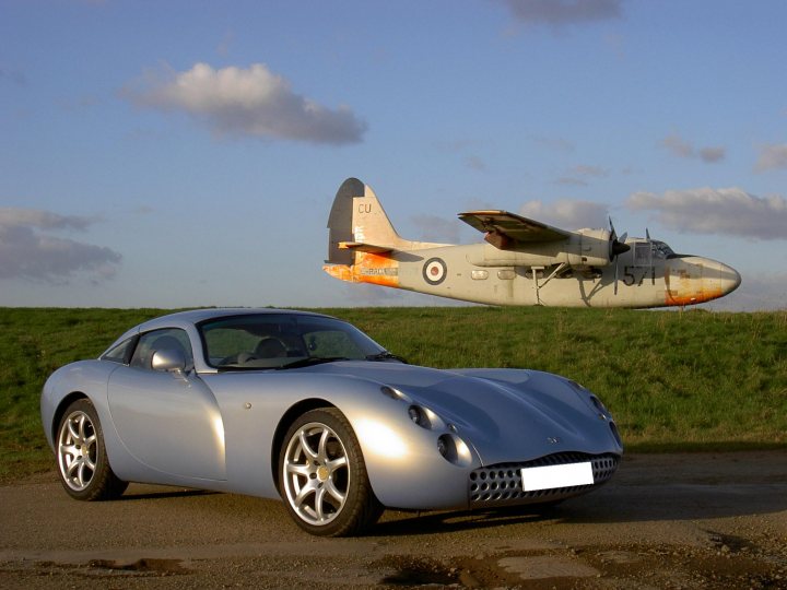 Your Best Tuscan Pic... - Page 8 - Tuscan - PistonHeads - This image features a gray sports car parked on the side of a road with a rally-like setting under a partly cloudy sky. The sports car has a distinctive classic design with black and silver wheels. In the background, an old, orange-marked double-engine plane seems to be taxiing on a grassy strip, adding a nostalgic and adventurous atmosphere to the scene. The overall color tone of the image is warm and inviting, perhaps suggesting a pleasant day for traveling or racing. The composition of the photo suggests that the car and the plane are positioned close to each other, possibly indicating a meeting point or a formal event.