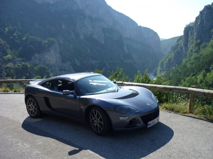 Pistonheads - The image depicts a sports car on a winding mountain road. It is a gray or silver color, and the car appears to be moving uphill on a clear day. The road is curved and there are steep drop-offs on the sides, revealing cliffs and a valley beyond. The car's sleek design contrasts with the rugged natural environment, creating a striking visual effect. The photo captures the car from a high angle, providing a sense of elevation in the landscape.