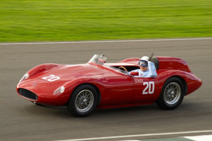 Revival - First Time and Excited - Page 1 - Goodwood Events - PistonHeads - A vintage red sports car is in focus, driving on a race track with a blurred speed level indicated with the number '20'. It has visible numbers '20', '20', and '20' on all three visible windows and the side of the car. The driver, also dressed in a white racing suit with blue goggles, is holding a red steering wheel. The background is a blurred green race track scene.