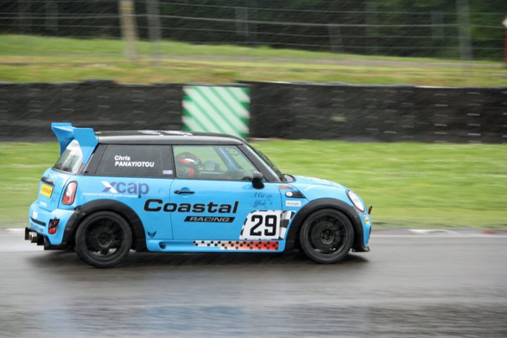 Pistonheads - The image features a blue Mini Cooper racing on a track. The car's race number is 29, and it bears the logos of Coastal Racing. The photo was taken in the midst of rain, as evident by the wet track and weather-beaten elements around the car. The setting is a natural outdoor environment with green hills and a clear sky.