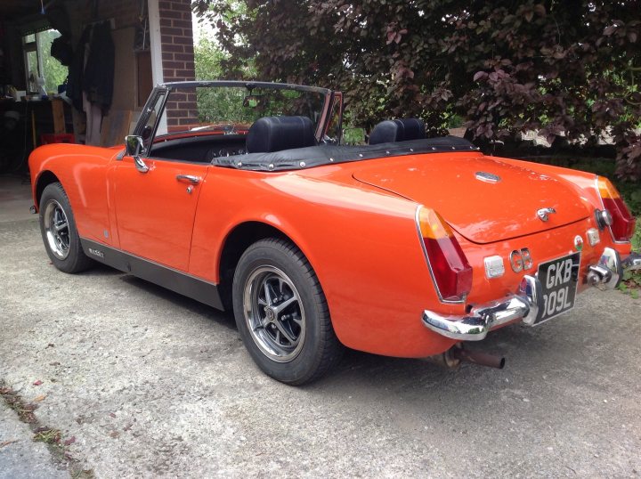 Midget Resto - Page 1 - MG - PistonHeads - This image shows a vibrant orange sports car parked on a concrete surface. The car has a distinctive black soft top and a black soft top backlight. It features classic white wall tires typical of vintage or classic cars. The vehicle has two doors and is equipped with a European license plate. In the background, there's a glimpse into a residential area, suggesting the car is parked on the street in front of a residential building.