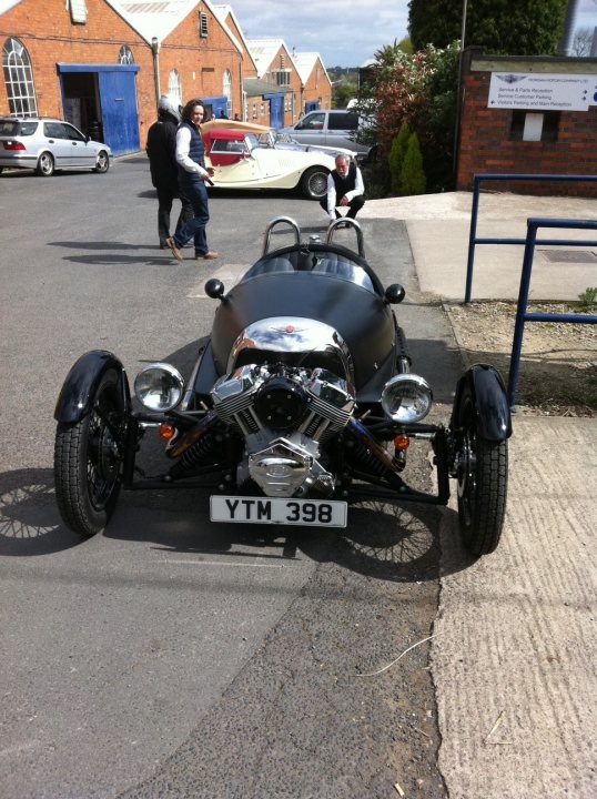 - Page 552 - General Gassing - PistonHeads - The image features an outdoor parking area of a small building. Dominating the scene is a unique three-wheeled motorcycle, with a prominent license plate that reads "YTM 398." The motorcycle is parked on a concrete surface and is well-lit, indicating bright sunshine. In the background, there are two people walking away, one of whom is carrying a handbag. A blue railing runs along the pavement next to the motorcycle, and there are some cars parked in the distance. The building has a red brick facade, and there's a blue door that partially matches the railing's color. The overall atmosphere suggests a normal day with good weather.