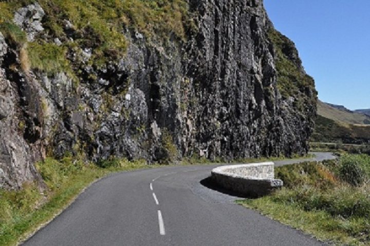 S Club Euro Tour 2015 - Le Tour de Franse - Page 15 - S Series - PistonHeads - The image shows a vehicle on a winding mountain road that appears to be carved into a cliff face. The cliff is steep and covered in moss, and the road is surrounded by natural vegetation. Above, the sky is clear, suggesting the photo was taken on a bright day with no impending weather. There is no visible text or writing in the image.