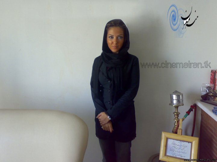 In the image, a woman stands in a room with a simple, clutter-free appearance. She is facing the camera with a slight smile on her face. The woman is wearing a black scarf and a dark-colored coat. There is no text on the image, and only minimalistic decorations can be seen in the background. The overall ambiance of the photo is quite calm and minimalist.