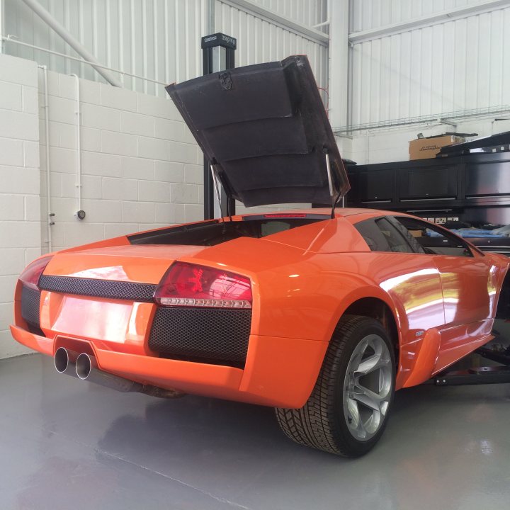 Update on Murcielago SG54..it's (almost) back - Page 1 - Diablo/Murcielago/Aventador - PistonHeads - The image showcases a striking orange or copper-colored sports car parked in a large industrial warehouse. The car appears to be a Lamborghini Huracan, known for its distinctive open-top design. An element that immediately catches the eye is the convertible roof, which is currently in the closed position. The garage itself is spacious and utilitarian, with hoses and what appears to be other machinery or vehicles visible in the background. The lighting suggests an indoor setting, likely a workshop or personal garage. There is no activity, suggesting that the car is stationary. The overall scene conveys a sense of luxury, enclosed in an everyday, functional environment.