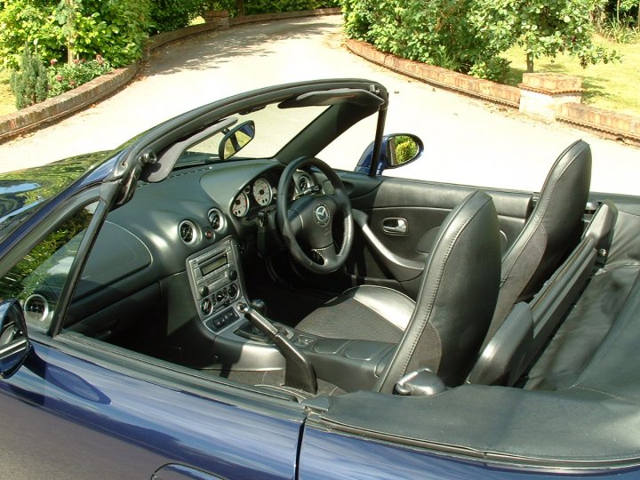 Pistonheads - The image features the interior of a luxury convertible car with the top down. The vehicle appears to be a two-door model and is seen through the rear passenger window. The steering wheel is on the left side, indicating that this might be a right-hand drive car. The dashboard is well-lit, suggesting either daytime or the use of ambient lighting inside the car. The car is parked on a road followed by a curb and green shrubbery, suggesting a suburban environment.