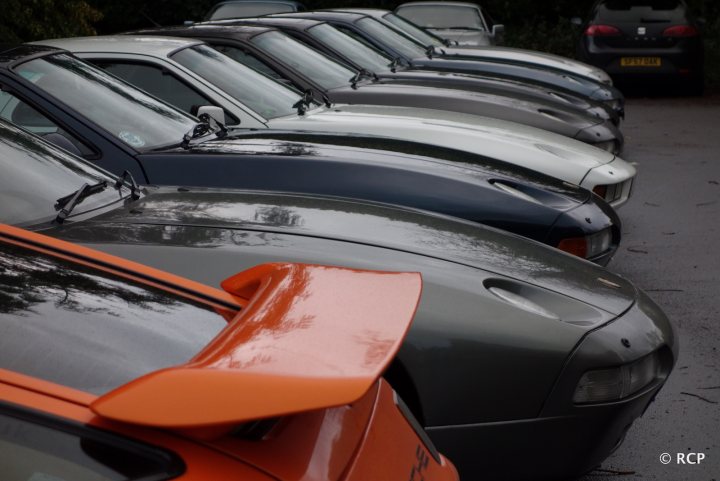 928 GT - worth? - Page 1 - Front Engined Porsches - PistonHeads - The image depicts a line of classic sports cars parked closely together. These cars are different models and are parked in an orderly manner looking toward the viewer. The hood of the orange car in the foreground is propped up, revealing its engine and interior. In the background, a modern car is partially visible, suggesting this line of classic cars is part of a mixed-themed parking lot.