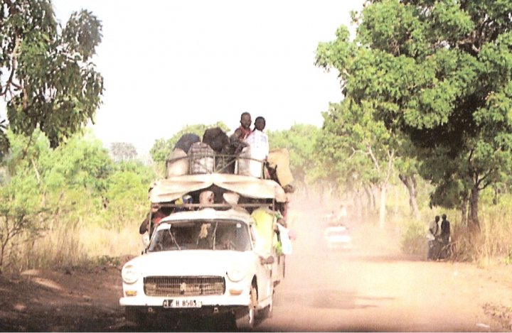 Peugeot 504 - Any still around? - Page 3 - Classic Cars and Yesterday's Heroes - PistonHeads - The image shows a scene from rural West Africa, featuring a group of people traveling on the back of a pickup truck. The truck is old and rusty, suggesting it has withstood many similar journeys. It is carrying several people with luggage, indicating a long journey. The road they are on seems rough and unpaved, adding to the sense of travel in an area with little infrastructure. The surroundings are covered in green trees and shrubs, typical of a savanna-like environment, with a light hazy sky above them. There is also a motorcycle visible in the background, further emphasizing the mode of transportation common in these regions.