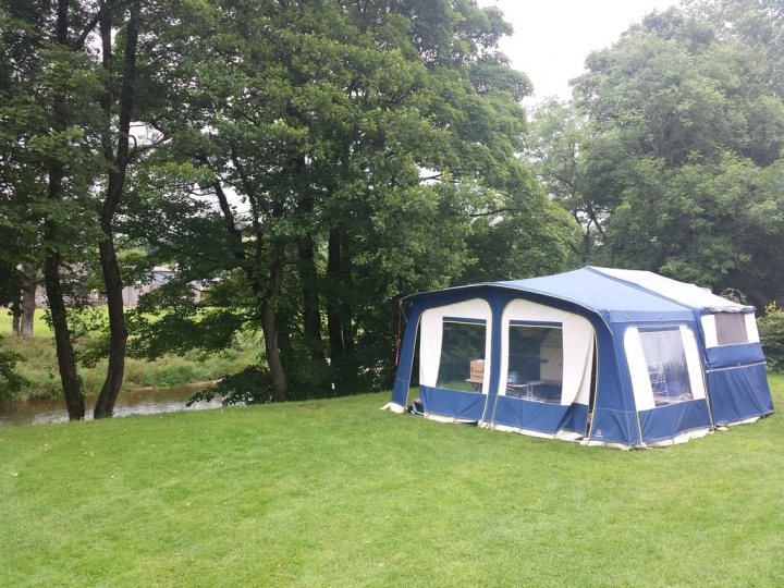 Camping for Pen Y Fan - Page 1 - Holidays & Travel - PistonHeads - The image captures a serene scene of a camper tent set up on a grassy field. The tent is primarily blue, with a white roof providing a stark contrast. A white door and three windows on the side suggest it's ready to accommodate future campers. The tent is pitched in a clearing that looks tranquil, surrounded by lush green trees and a small river with a gentle flow. The sky overhead is filled with clouds, adding a subtle mood to the setting.