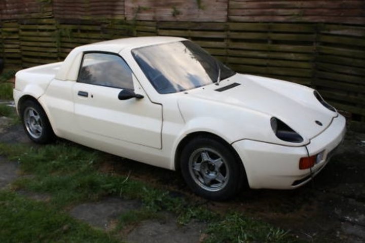 Evening Gtm Rossa Ebay Pistonheads Ends - The image shows a compact, white two-door vehicle parked on grass. The car has unique, skinny headlights that narrow towards the front, giving it a distinct look. It appears to be an older model, as indicated by the style of the wheels and the lack of modern features visible in the photograph. The background features a fence, partially obscured by the car, suggesting a rural or suburban setting. The lighting suggests it is either evening or the car itself is casting a shadow, indicating that the photo was taken during daylight.