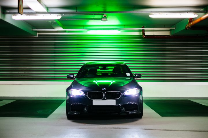 Is the f10 M5 a fun car? - Page 2 - M Power - PistonHeads - This image features a BMW car parked in an underground parking garage. The car, which is black, is positioned centrally in the frame with its headlights on. The backdrop consists of green neon lighting naturally reflecting off the garage walls and a gray painted floor. The overall lighting gives the scene a cool contrast.