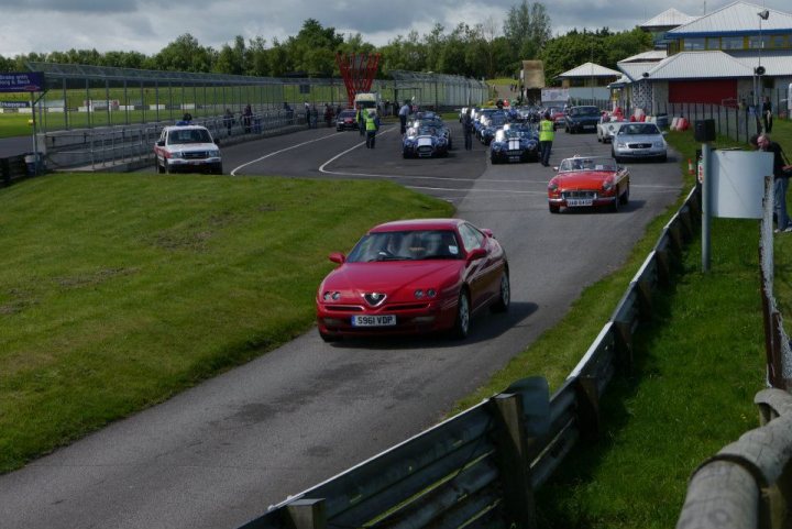 A red car is driving down the street - Pistonheads - In this dynamic image, a red Alpine sports car is the center of attention, speeding away from the viewer down a road alongside a wooden fence. The car is passing a silver Prius, leaving it in its wake. This scene takes place in a spacious parking lot that's adjacent to a grassy area dotted with leafless trees. In the distance, a fence runs parallel to the road, adding depth to the image. There's also a red building visible, which contrasts with the natural elements of the scene. The cloudy sky suggests that this exciting chase might be taking place under a grey, overcast sky.