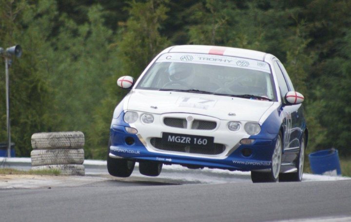 Pictures of your racecar thread - Page 1 - UK Club Motorsport - PistonHeads - The image shows a white and blue rally car in an outdoor setting, likely a race track, with greenery in the background. The vehicle is in motion, slightly tilted to the right side of the frame, implying a course turn. The rear tire is lifting off the ground, indicating a moderate speed. There is a car number visible, but specific details like the make, model, or brand are not entirely clear. The image appears to capture the dynamic nature of a rally or track race.