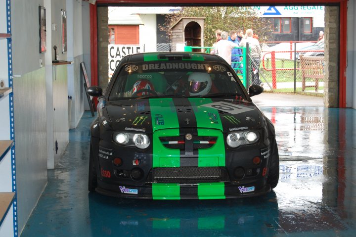 Stroke Association Supercar Day Castle Pistonheads Combe - The image shows a black sports car with vibrant green and black stripes running down its side, parked indoors under a building. The vehicle's design is sleek and low-to-the-ground, indicative of a high-performance model. There is a clear reflection of the car in the surrounding puddle, showing its mirror image. Adjacent to the car, a garage door has an open vertical position, inviting the viewer into the rest of the indoor space. From the reflection and the background, it seems to be raining outside the garage.