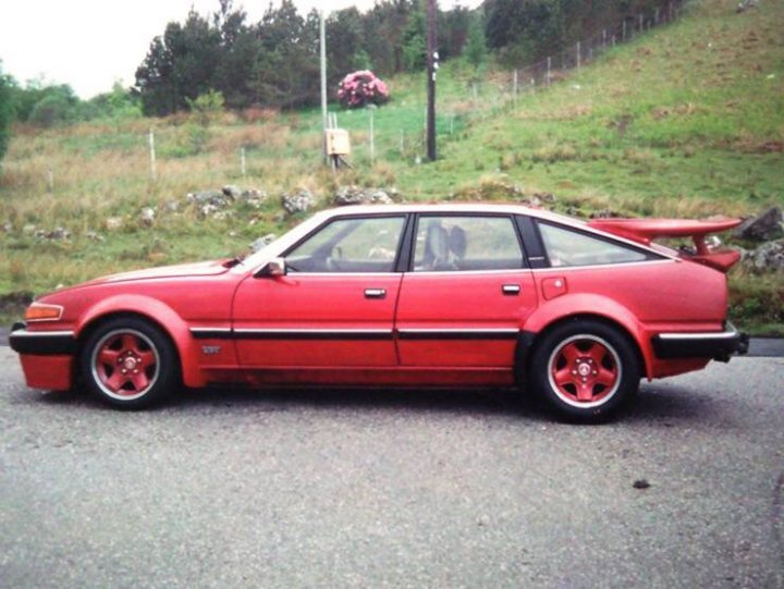 1985 Rover 3500 Vitesse - Page 16 - Readers' Cars - PistonHeads - The image features a vibrant red sports car parked on the side of a road. This is not your average sedan; it's an eye-catching vehicle with a sleek body, long hood, and short tail. A distinctive feature is its rear spoiler, adding to its sporty aesthetic. The wheels are silver, providing a nice contrast to the red body. A person can be seen in the background, but their identity or activity is not clear. The car seems well-maintained, indicating frequent use. The overall scene suggests a sunny day with pleasant weather, making it a perfect setting for a drive.