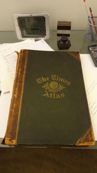 Something different...1895 Times Atlas of the World - Page 1 - Books and Literature - PistonHeads - The image showcases a weathered green book lying open on a desk, with the title "The Times Atlas" prominently displayed. The book appears to be old and well-read, indicative of frequent use. Next to the book, there's a white digital thermometer and a calculator, suggesting that the desk belongs to someone involved in financial or scientific work. The photograph might imply that the owner of the items is engaged in reading, calculation, or planning related to geography.