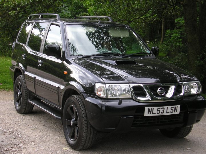 tuned terrano 3.0 di sve - Page 1 - Readers' Cars - PistonHeads - The image shows a black Nissan 4x4 parked on a gravel road. The vehicle has a sunroof, black alloy wheels, and a front licence plate that reads "NL53 LSN." It's situated in a lush green environment with trees in the background, hinting at a wooded area. The sky can be seen through the vehicle's open sunroof, suggesting a time of day when the weather is clear. The setting appears calm and peaceful.