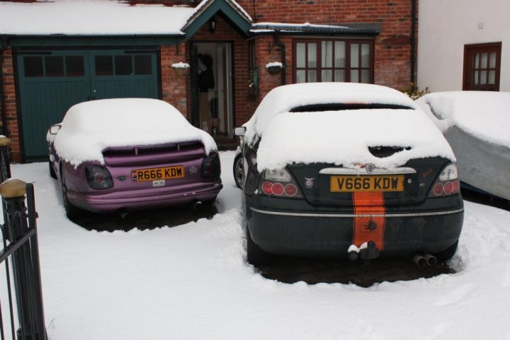 Show us your REAR END! - Page 120 - Readers' Cars - PistonHeads - The image portrays a peaceful winter scene with two cars parked by a red brick building. The car closest to the viewer is covered with a thick layer of snow, suggesting that it hasn't been driven for quite some time. Its license plate is clearly visible against the white snowy background. The second car is parked slightly farther away and is partially obscured by the first car due to the camera angle. The cars appear to be secured with snow chains, a common precaution during snowy conditions. The brick building provides a contrasting structure against the snowy backdrop.