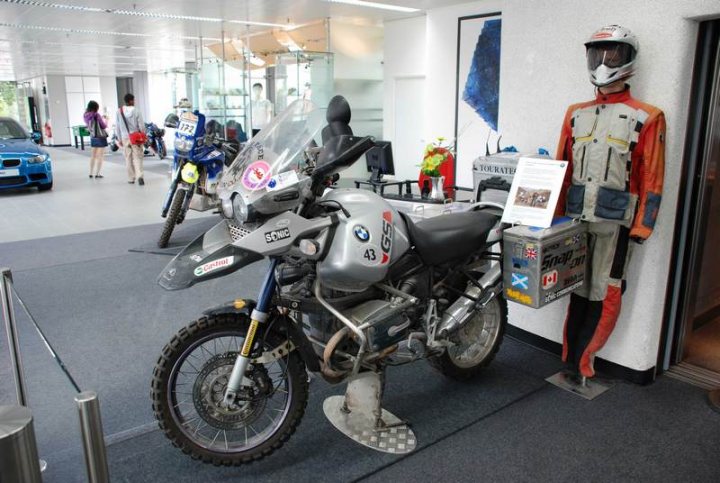 Pistonheads Service Sunday - The image shows an indoor motorcycle and scooter exhibition, likely in a dealership or museum-like setting. The centerpiece of the scene is a silver BMW motorcycle with a protective windshield and a prominent "SCOWR" sticker on its front. The motorcycle is stationed on a gray carpet.

The showroom itself is modern and brightly lit, with windows allowing natural light to enter. In the background, a row of motorcycles and scooters are visible, suggesting the venue is dedicated to such vehicles. A mannequin dressed in a racing suit is situated to the right, partially visible, while another figure dressed in an orange and beige outfit is displayed to the left.

Various decorative items such as potted plants and a vase with a blue flower can be seen in the background, adding a decorative touch to the space. The presence of people in the background, potentially customers or visitors, adds a sense of scale to the scene. The environment suggests an open and engaging atmosphere for motorcycle enthusiasts.