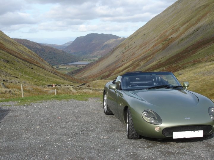 Help identify this Lake District location? - Page 1 - General Gassing - PistonHeads - The image showcases a vibrant green Mazda Miata sports car parked on a gravel road in a mountainous area. The car is positioned such that the angle from the rear to the front of the vehicle is highlighted, allowing for a clear view of its sleek design. The surrounding landscape is characterized by grassy fields and majestic mountains, with a serene lake visible in the distance. Above, the sky is a clear blue with a few clouds scattered across it, creating a serene and picturesque backdrop for the scene. This setting suggests a journey through scenic and natural beauty, reinforcing the adventurous spirit that the Miata represents.