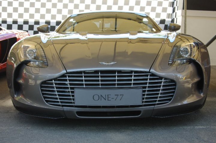 Anyone been to the Moving Motorshow before? - Page 1 - Goodwood Events - PistonHeads - The image shows a silver sports car with a front license plate that reads "One-77". The car appears to be a sleek, elegant vehicle with a modern design, showcasing its luxurious features such as the grille and the headlights. The car is parked, as it is turned slightly towards its right, and there is a black and white checkered pattern on the background, which is a typical sight at race tracks, adding to the sense of speed and performance this car offers. The overall impression is of a high-end automobile placed in a context that emphasizes its status and design.