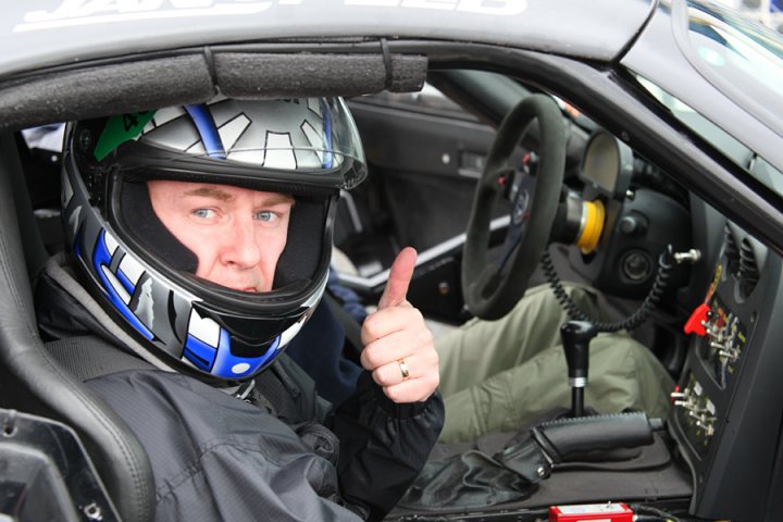 Pistonheads - The image shows a person wearing a motorcycle helmet, making a thumbs-up gesture. They are sitting in the driver's seat of a race car. The person has a confident expression on their face. In the background, there is another person partially visible, suggesting a shared activity. The race car appears to be in motion with various controls and sensors around the driver.