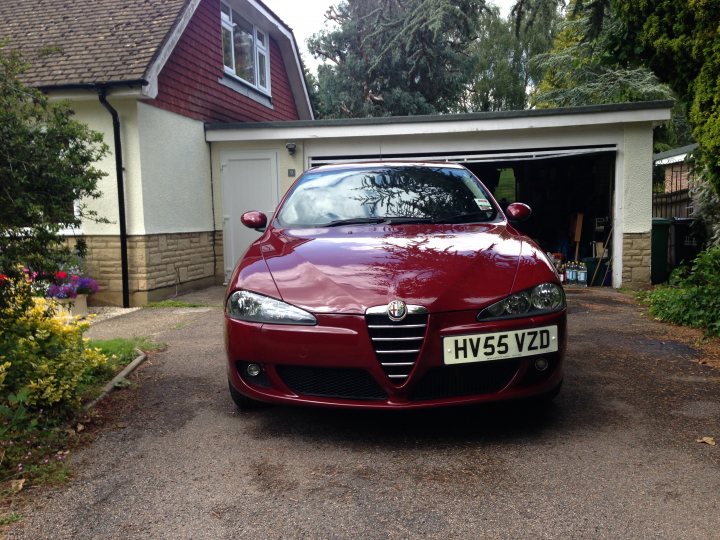 Alfa 147 would you ?  - Page 1 - Alfa Romeo, Fiat & Lancia - PistonHeads - The image portrays a distinct scene featuring a glossy red Audi parked in front of a detached garage. The garage features a roller shutter and a single white door. The driveway is lined with a variety of plants, including flowers that add a touch of color to the otherwise neutral tones of the scene. The overall setting suggests a domestic environment, possibly during a weekend or resting day.