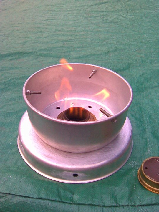 Trangia or Primus stove? - Page 1 - Tents, Caravans & Motorhomes - PistonHeads - A small metal exhaust hood is seen, with a flame above it on a fabric surface. The hood has a silver-colored rim with two small perforations, possibly for ventilation or mounting purposes. A round hole is in the center, where the flame is exhibited. The fabric below and nearby the burner appears green. Lastly, a circular object with a metallic core and brown outer border is casually placed in the bottom right corner of the image.