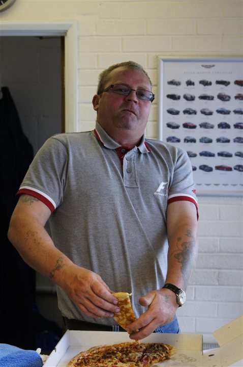 Work Halls Pistonheads Tony Experience - This is a straightforward photo of a man preparing to eat from a pizza box. He's wearing a gray short-sleeve shirt with an accent on the sleeve and a red tie, indicating a semi-formal dress code. The man is holding a slice of pizza and looking directly at the camera, giving the photo a candid feel. It's a moment captured during a mealtime or possibly a break, as there is a clock in the background that seems to indicate the time. The pizza itself looks deliciously topped with cheese and what appears to be vegetables, but the specifics aren't clear from this angle. The overall setting seems to be a casual dining environment.