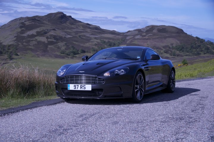 Some pics from a recent trip to the Scottish highlands.... - Page 1 - Aston Martin - PistonHeads - The image showcases a luxurious black sports car parked on the side of a highway. The car is set against a backdrop of sheer rock cliffs, which provide a dramatic and contrasting landscape. The perspective of the car in the foreground, with the cliffs in the background, gives a sense of depth and scale. The sky above appears to be a clear day, casting a pleasant light on the scene. The car is positioned angled to the camera, allowing for a comprehensive view of its design and features.