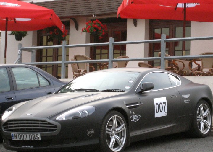 How about an Aston photo thread! - Page 11 - Aston Martin - PistonHeads - The image features a black Aston Martin parked outside a white building. The car sports a license plate reading "DP07", and there's a distinctive James Bond 007 logo visible. Additional elements include red umbrellas and potted plants, with another car parked nearby. The scene suggests a calm residential area.