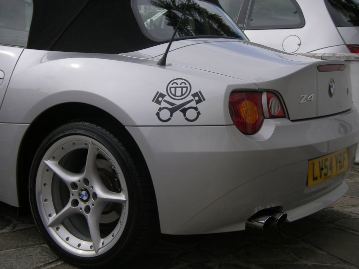 Pistonheads - The image features a silver BMW car parked on a cobblestone road. The car is viewed from the back and has a distinctive silver and black circular logo on its trunk, depicting a smiley face with a camcorder. The tail lights and license plate are also visible. There are other vehicles in the background, one of which is a large commercial vehicle.