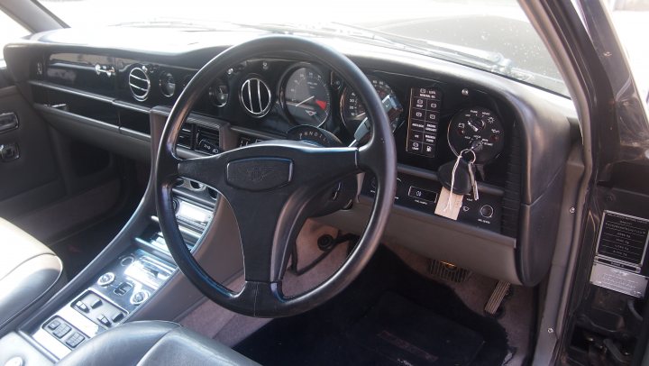 Have we been foolish? Yes but we're all grinning from ear to - Page 1 - Rolls Royce & Bentley - PistonHeads - The image displays the interior view of a car, specifically the driver's seat area. A black leather steering wheel, equipped with various buttons and controls, is at the center of the view. In front of the steering wheel, there's a dashboard with various gauges and indicator lights. To the right of the steering wheel, there is a gear shift. The overall color scheme of the interior is a combination of black and silver.