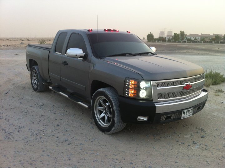 Not what  I had in mind but ....... - Page 1 - Yank Motors - PistonHeads - The image shows a dark-colored pickup truck parked on a flat, open surface, which appears to be a sandy area. The vehicle has a prominent front bumper and grille design, with fog lights integrated into the grille. The truck is equipped with five-spoke off-road wheels, and there is a visible license plate at the front. In the background, there's a hint of a clear sky and a sparse landscape with minimal vegetation and buildings at a distance. The lighting suggests it might be late afternoon or early evening, with natural light still illuminating the scene.