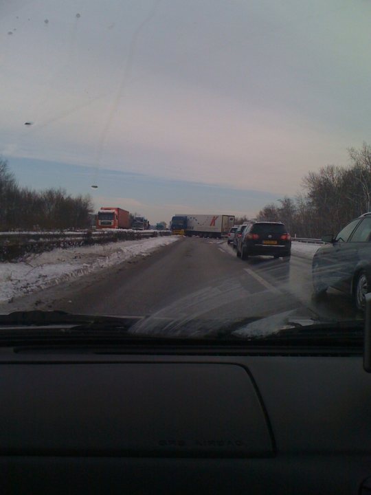 Pistonheads - The image captures a daytime scene on a highway. Beneath the car windshield, the view shows a major road or highway. There is snow on the ground, indicating cold weather. Several vehicles including cars and a bus are visible, some stationary and others appearing to be moving. Notably, a truck is sitting idle on the side of the road, perhaps awaiting clearance or perhaps experiencing a breakdown. The sky overhead is overcast, adding to the impression of a cold and possibly recent snow storm.