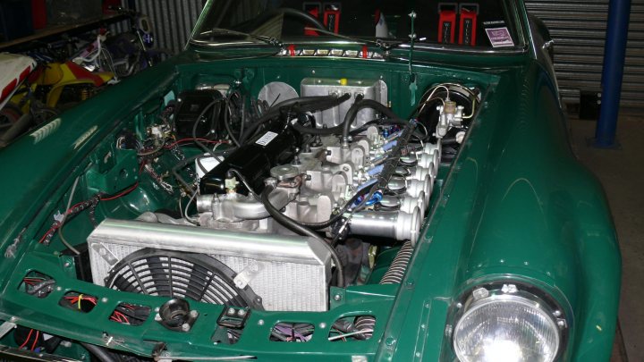 MGC gets new engine - Page 1 - MG - PistonHeads - This image presents a close-up view of the engine bay of an old, green car, showing the intricate details of its mechanical components. The engine is the main focus, with various wires and cables visible, along with the shiny metal surfaces of the engine cover and other parts. Surrounding the engine, you can see other components of the car, such as the radiator, which is a key element in the engine cooling system. The overall scene suggests a garage or workshop setting, where the car might be undergoing maintenance or repair.