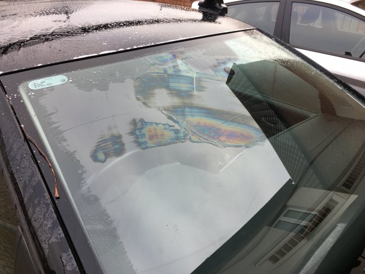 How to get grease off a windscreen? - Page 1 - Bodywork & Detailing - PistonHeads - The image shows the front windshield of a vehicle, reflecting the backdrop. The scene is mainly black and white, indicating an overcast or cloudy day. There is a visible reflection of a building's windows on the hood of the car. Additionally, the styrofoam exterior of the car seems to be dirty or wet, likely from rain or snow. There are also reflections visible on the car's windows, which add to the image's overall muddiness aesthetic. The image is a photograph taken in the daytime.