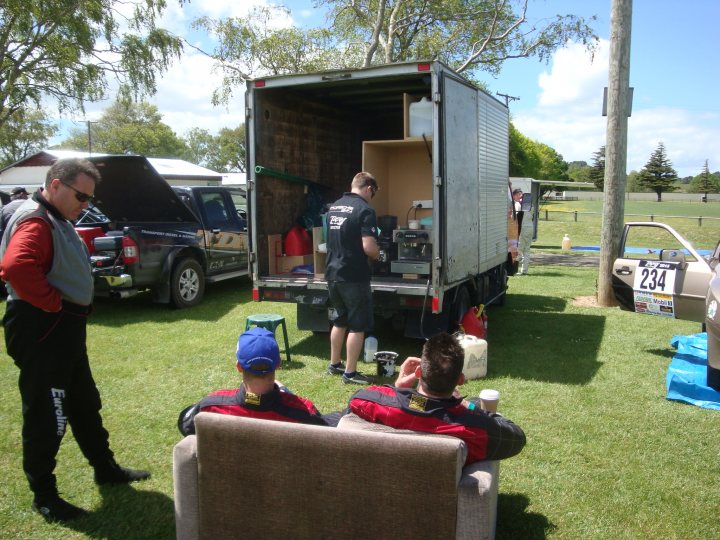 Targa 2013 - Page 1 - New Zealand - PistonHeads - The image captures a casual, social scene in an outdoor setting. A man is standing next to an open truck, inspecting a wooden pallet. Nearby, two other individuals are sitting on a couch and enjoying the sunny weather, a testament to the relaxed and leisurely atmosphere. A car is parked, and personal belongings such as a suitcase and a skateboard are scattered around the area. The overall ambiance suggests a communal gathering or a meet-up of friends or colleagues.