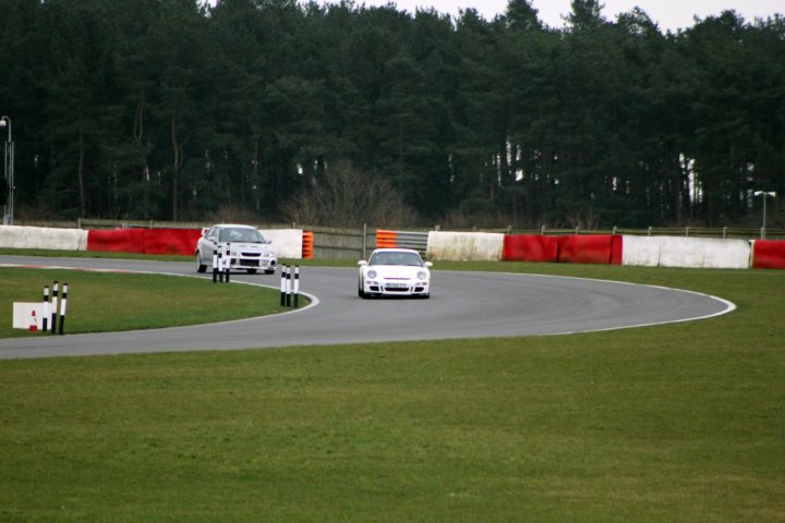 Track day at Snetterton Friday 15th February - Page 1 - East Anglia - PistonHeads - The image depicts an action-packed scene on a race track. Two cars, one white and the other black, are speeding down the track, taking the corners simultaneously. They are situated on the right side of the track. In the background, there are palm trees and a forest of pine trees, adding a tropical or Southern vibe to the setting. The entire scene is a beautiful blend of speed, competition, and natural beauty.