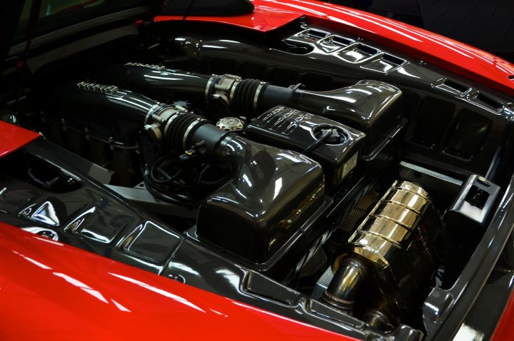 16M - Page 1 - Ferrari V8 - PistonHeads - The image displays the underside of a sleek red sports car, revealing its engine compartment. The engine is equipped with a black air intake filter, which is positioned in the center. In the background, the silhouette of a clock is visible, suggesting that the photo might have been taken in a setting with a clock present.