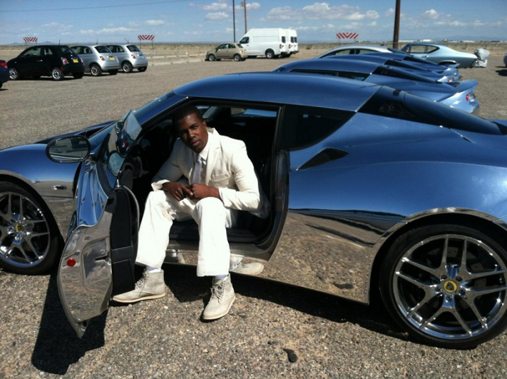 A man standing next to a car on a street - Pistonheads - In the image, a man is seated in the driver's seat of a highly reflective, long silver convertible sports car parked in a gravel parking area. The man appears to be waiting or relaxing, dressed in a white suit and mustard yellow shoes. The car's hood is raised, and the overall scene suggests a vintage or classic automobile. The background shows a vast, empty parking lot with a clear sky and a few other vehicles parked in the distance.