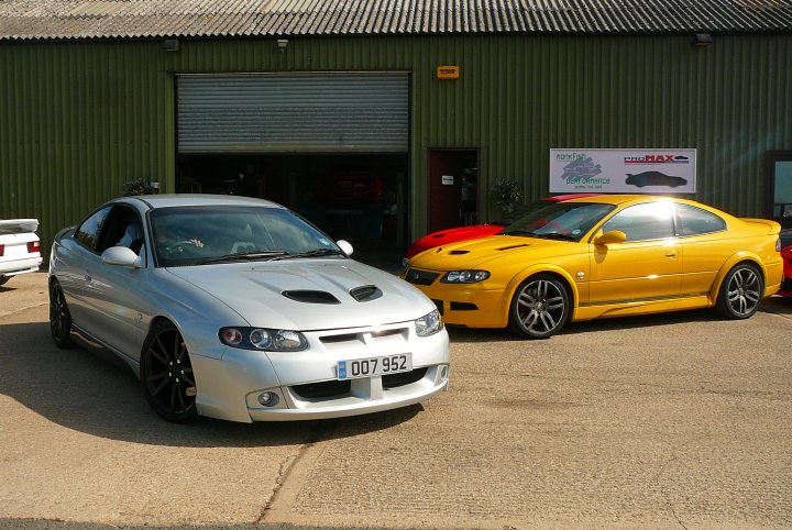 Pistonheads - The image shows an outdoor setting with two cars parked on a tarmac area. The car on the left has a customized exterior with a necessity symbol on the side and is clad in a metallic silver finish. The car on the right is a vibrant yellow color with a sleek design, both cars sporting large alloy wheels. Behind the cars, there is a green building with a roller shutter door, and on the side of the building, a sign indicating a workshop or business establishment is partially visible.