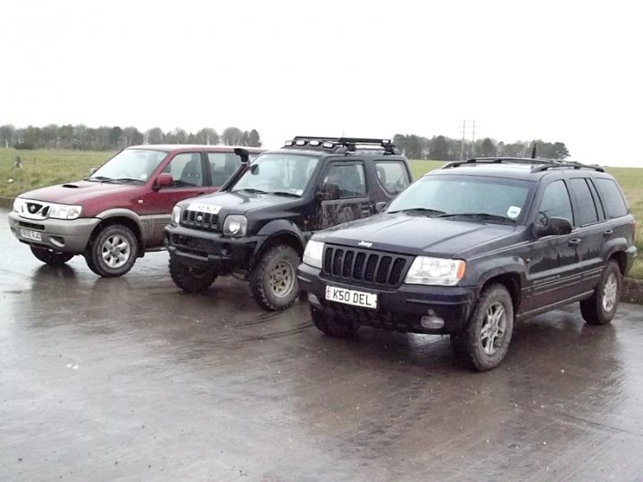 Trip onto the Plain in the next few days? - Page 1 - Off Road - PistonHeads - The image shows three Jeep Wrangler vehicles parked side by side on a muddy lot. The leftmost vehicle appears to be a red or burgundy model, possibly missing a front wheel, with the hood down and the back door open. The center vehicle is black and has a spare tire mounted on the back door. The rightmost vehicle is grayish and looks unmodified compared to the other two. In the background, there is a field and a row of trees, suggesting a rural or semi-rural setting.