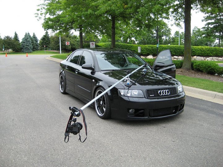 Car Camera Rigs - Page 1 - Photography & Video - PistonHeads - The image shows a scene in a parking lot where a black Audi car is parked next to a grassy area. A tripod is stuck in the ground near the car, with the head of a camera attached to it. The camera is pointed towards the car, suggesting that it might be set up for a photography session or a video recording. The background features other trees and a green field, giving the setting a natural and serene atmosphere.