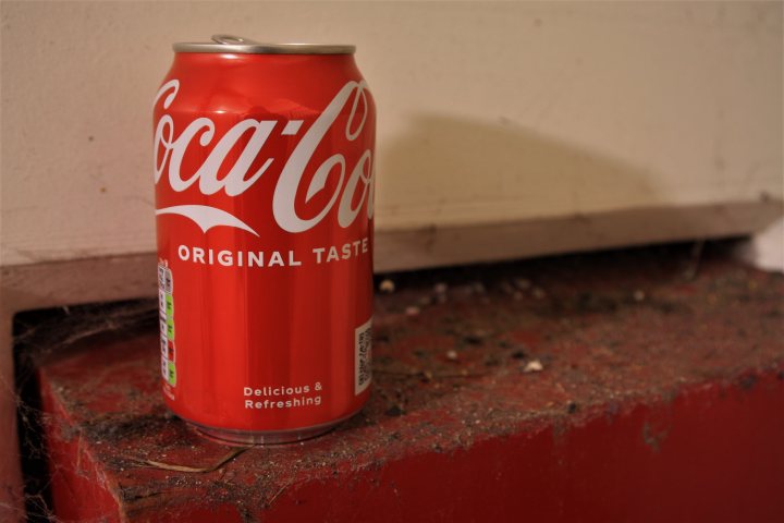 Pistonheads - The image shows a can of Coca-Cola with the text "Original Taste" prominently displayed. It's leaning against what appears to be a wooden surface, possibly part of a wall or bench. The background is dark and out of focus, which puts the emphasis on the soda can. There are no people or other objects in the immediate vicinity, making the can the central subject of this image.