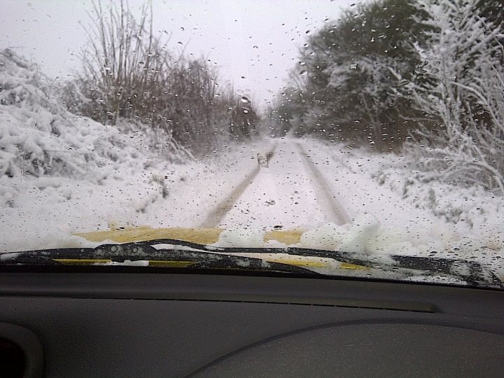 Commuting from Bath to Portbury - Page 1 - South West - PistonHeads - The image depicts a winter scene through the windshield of a vehicle. The windshield is covered in snow and ice, which is beginning to melt, creating streaks on the glass. The road outside is also covered in snow, and it seems to be a rural or forested area, as there are trees visible on either side of the road. The weather appears overcast, and the visibility is reduced due to the inclement conditions. Despite the challenging driving conditions, there is a sense of tranquility in the otherwise harsh winter weather.